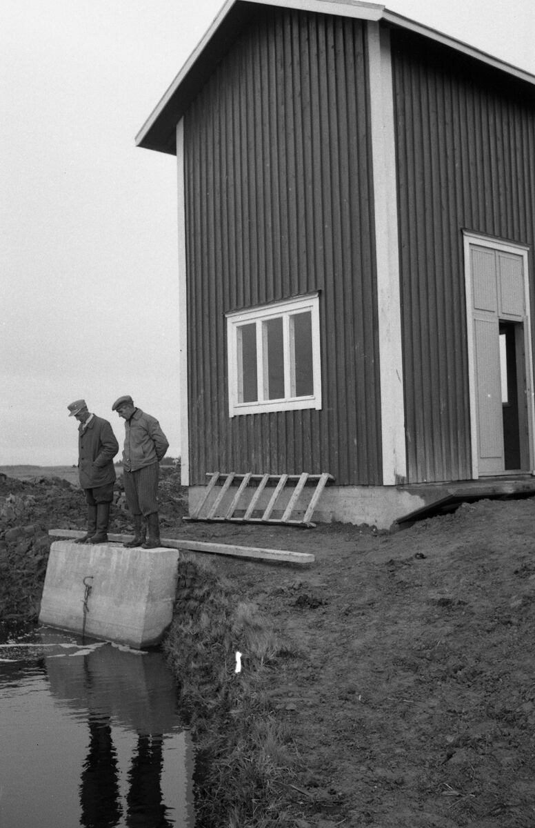 ???resource.thumbnail???: Två män betraktar vattennivån utanför ett pumphus.